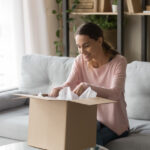 Happy woman sitting on couch at home opening carton box received parcel package from relatives or shopper make order internet website satisfied client, easy and fast service commerce delivery concept